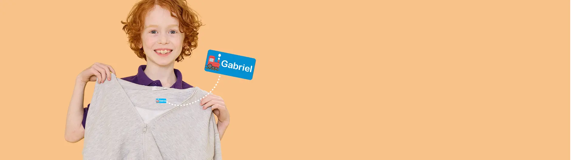 Menino a segurar um casaco cinzento-claro com fecho e uma etiqueta de nome azul com comboio e o nome Gabriel na etiqueta de lavagem